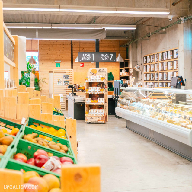 L'intérieur du magasin d'alimentation naturelle Le Bon Wagon à Eupen, avec des étagères en bois contenant des fruits et légumes dans des caisses vertes à gauche, un comptoir réfrigéré avec des produits alimentaires à droite, et deux caisses enregistreuses au fond avec des panneaux indiquant KASSE 1 et KASSE 2.