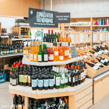 Une section du magasin d'alimentation naturelle Le Bon Wagon à Eupen, avec des étagères remplies de bouteilles de vin et d'autres boissons alcoolisées. Il y a un panneau indiquant CONGÉLATEUR TIEFKÜHLWARE et un autre panneau indiquant WEIN DES MONATS VIN DU MOIS. Les bouteilles sont organisées par type et couleur, avec des vins rouges, blancs et rosés visibles.