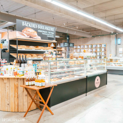 Une boulangerie artisanale à l'intérieur du magasin d'alimentation naturelle Le Bon Wagon à Eupen. Il y a des étagères remplies de pains et de viennoiseries, ainsi qu'une vitrine réfrigérée contenant divers produits de boulangerie. Un panneau au-dessus des étagères indique Bäckerei-Boulangerie en plusieurs langues, y compris Handgemacht et Fait Main. Il y a également une petite table avec des bouteilles de boissons en vente.