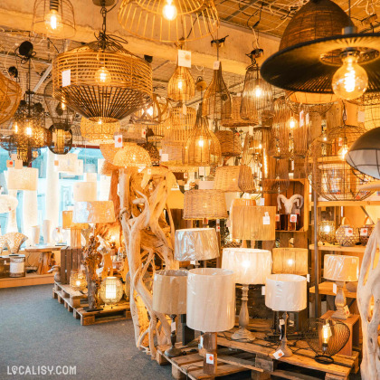 L'intérieur du magasin de luminaires Mabrilux à Welkenraedt, avec une grande variété de lampes suspendues et de lampes de table, principalement en matériaux naturels comme le rotin et le bois. Les luminaires sont disposés sur des palettes en bois et accrochés au plafond, créant une ambiance chaleureuse et accueillante. Les étiquettes de prix sont visibles sur plusieurs articles.