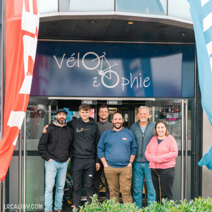 Devanture du magasin de vélo Velozophie à Welkenraedt. Un groupe de six personnes debout devant l'entrée du magasin. L'enseigne du magasin est bleue avec le nom Vélozophie écrit en blanc, où le O est stylisé comme une roue de vélo. Des drapeaux colorés sont visibles de chaque côté de l'entrée.