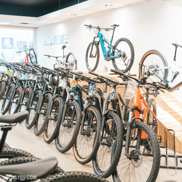 L'intérieur du magasin de vélos Velozophie à Welkenraedt, avec une rangée de vélos de montagne alignés sur le sol, ainsi que quelques vélos suspendus sur un présentoir au mur. Le logo CUBE est visible sur le mur à l'arrière-plan. Les vélos sont de différentes couleurs et modèles, et l'éclairage du magasin met en valeur les détails des vélos.