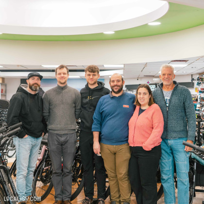 Un groupe de six personnes debout dans le magasin de vélos Velozophie à Welkenraedt. Ils sont entourés de vélos et d'accessoires de vélo. Le magasin a un intérieur bien éclairé avec des lumières au plafond et des étagères remplies de produits cyclistes en arrière-plan.