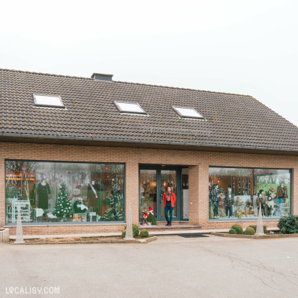 Devanture du magasin de vêtements pour enfants et bébés Ideal Baby & Junior Company à Waimes. Le bâtiment a un toit en tuiles avec trois fenêtres de toit. La façade est en briques et comporte deux grandes vitrines de chaque côté de l'entrée principale. Les vitrines sont décorées avec des articles de Noël, y compris des sapins de Noël, des mannequins habillés et d'autres décorations festives. Une personne se tient à l'entrée du magasin.