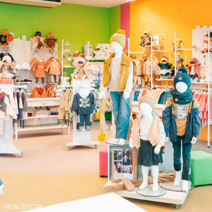 L'intérieur du magasin de vêtements pour enfants et bébés Ideal Baby & Junior Company à Waimes, avec des murs verts et orange, et il est bien organisé avec des vêtements pour enfants exposés sur des mannequins et des étagères. Les mannequins portent des tenues variées, y compris des vestes, des bonnets, des écharpes, des jupes et des jeans. Il y a également des vêtements suspendus sur des cintres et des accessoires comme des chapeaux et des chaussures.