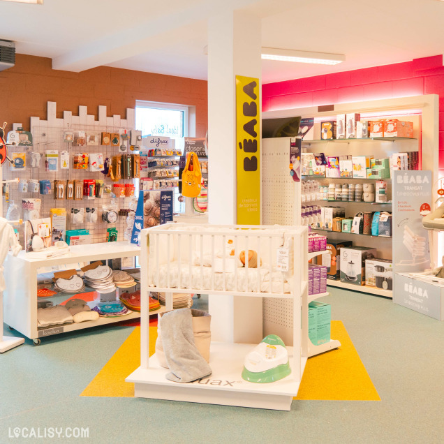 L'intérieur du magasin de vêtements pour enfants et bébés Ideal Baby & Junior Company à Waimes, avec divers produits pour bébés et jeunes enfants, tels que des accessoires de puériculture, des jouets, des vêtements et des articles de soin. Au centre de l'image, il y a un lit de bébé blanc avec des articles de literie et des jouets. À gauche, il y a des étagères et des présentoirs avec des produits suspendus, tandis qu'à droite, il y a des étagères avec des boîtes et des paquets de produits.