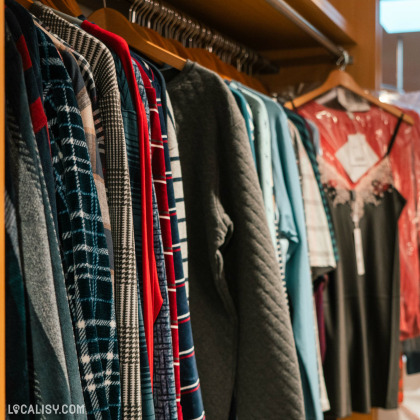 Une rangée de vêtements suspendus sur des cintres dans le magasin de lingerie Lingerie Huppertz à Waimes. Les vêtements incluent des chemises à carreaux, des pulls, et une robe noire avec des détails en dentelle. Le magasin semble bien organisé avec une variété de styles et de motifs.