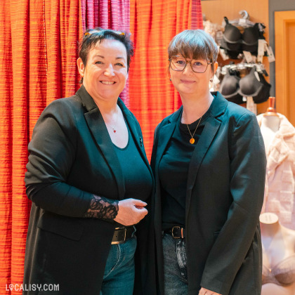 Deux personnes souriantes dans un cadre professionnel, vraisemblablement dans le magasin de lingerie Lingerie Huppertz à Waimes. À l'arrière-plan, on peut voir des articles de lingerie accrochés et une ambiance chaleureuse avec des rideaux rouges. Si cela représente le magasin Lingerie Huppertz à Waimes, l'image illustre probablement une partie de l'équipe ou les responsables, mettant en avant une atmosphère conviviale et accueillante.