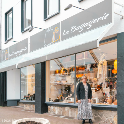 Devanture du magasin de maroquinerie La Bagagerie à Malmedy. La façade est moderne avec des auvents gris et des vitrines bien éclairées, exposant divers sacs et accessoires. Une personne se tient devant le magasin, habillée de manière élégante. Le logo du magasin, représentant des sacs, est visible sur les auvents.