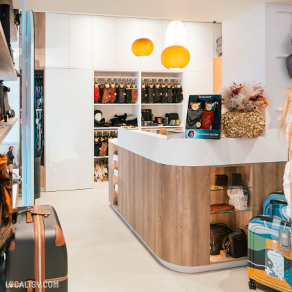 Un comptoir en bois et blanc avec des étagères intégrées contenant divers articles comme des sacs et des accessoires dans le magasin de maroquinerie La Bagagerie à Malmedy. Derrière le comptoir, il y a des étagères blanches remplies de gants de différentes couleurs et styles. Deux lampes suspendues orange éclairent la zone. À droite du comptoir, il y a une étagère avec des valises colorées. Un présentoir sur le comptoir montre des gants pour écrans tactiles.