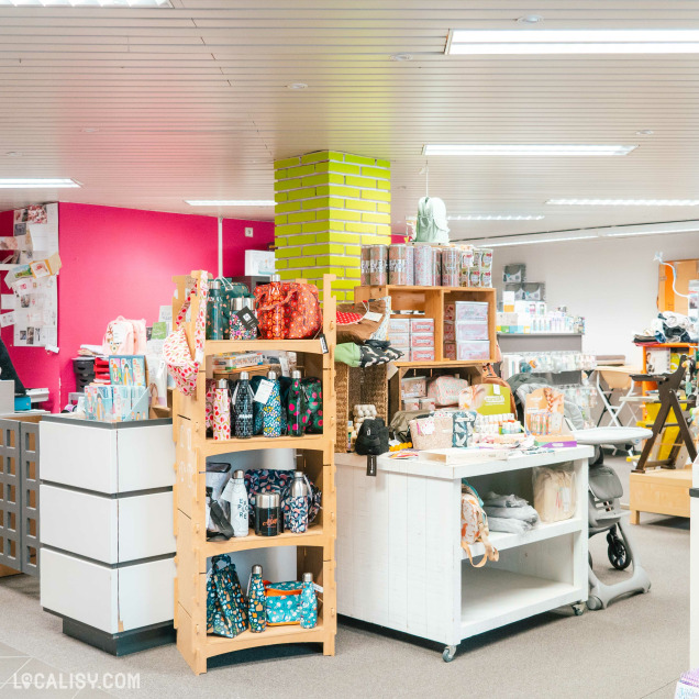 L'intérieur du magasin de produits d'occasion Toup'tibou à Waimes, avec plusieurs étagères en bois remplies de sacs colorés, de bouteilles et d'autres articles divers. Un pilier central est peint en jaune vif avec des briques apparentes. À l'arrière-plan, on peut voir un comptoir avec des articles supplémentaires et des étagères contenant des boîtes et des produits divers.