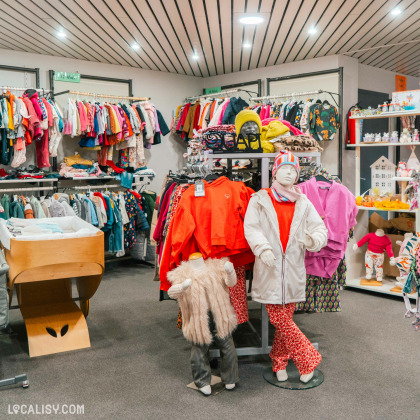 L'intérieur du magasin de produits d'occasion Toup'tibou à Waimes. On y voit plusieurs étagères et portants remplis de vêtements colorés pour enfants, allant des manteaux aux pulls en passant par des pantalons et des accessoires comme des bonnets. Au centre de l'image, il y a deux mannequins habillés avec des vêtements d'hiver, l'un portant un manteau blanc et l'autre une veste en fausse fourrure.