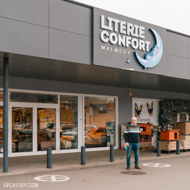 Devanture du magasin de literie Literie Confort à Malmedy. Le magasin a une enseigne avec le nom Literie Confort en grandes lettres blanches, accompagnée d'une illustration d'une personne allongée sur un croissant de lune. Devant le magasin, il y a un homme debout sur le trottoir. À travers les vitrines, on peut voir des lits et des matelas exposés à l'intérieur. À droite de l'entrée, il y a des articles empilés, probablement des meubles ou des accessoires de literie.