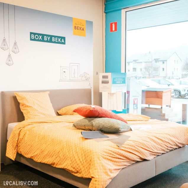 Un lit exposé dans le magasin de literie Literie Confort à Malmedy, avec une enseigne BOX BY BEKA et le logo BEKA visible sur le mur derrière le lit. Le lit est recouvert de draps jaunes avec des motifs, et plusieurs coussins de différentes couleurs sont posés dessus. À côté du lit, une affiche indique un prix de 1.800 €. À l'extérieur, on peut voir des voitures garées et des bâtiments à travers une grande fenêtre.