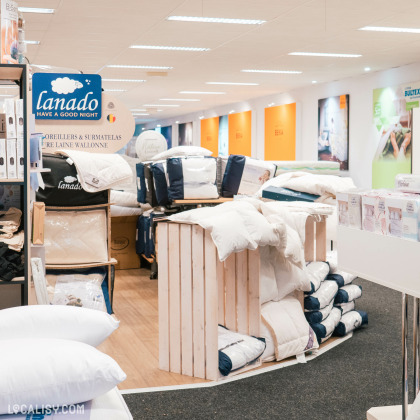 Des oreillers, des surmatelas et des couettes empilés et exposés sur des étagères en bois dans le magasin de literie Literie Confort à Malmedy. Un panneau indique la marque Lanado avec le slogan Have a good night. Le magasin est bien éclairé et montre un espace bien organisé avec des produits de literie de haute qualité.