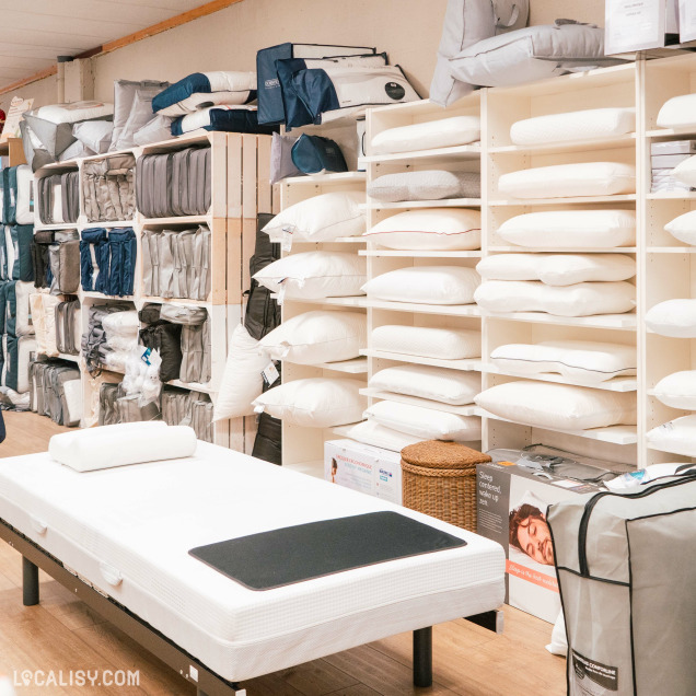 Un lit d'exposition au premier plan, entouré de nombreux oreillers et coussins disposés sur des étagères à droite dans le magasin de literie Literie Confort à Malmedy. À gauche, il y a des étagères remplies de housses de couette, de draps et d'autres articles de literie.