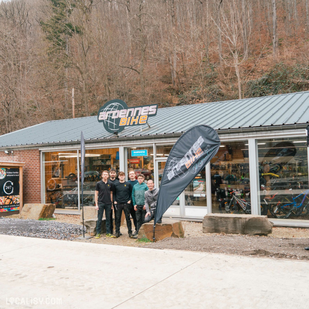 Devanture du magasin de vélos Ardennes Bike à Spa. Le magasin a une façade en verre avec un toit en métal. Devant le magasin, il y a un groupe de cinq personnes posant pour la photo. Il y a également un drapeau noir avec le logo Ardennes Bike planté dans le sol. À l'intérieur du magasin, on peut voir des vélos et des accessoires de vélo exposés. Le magasin est situé dans une zone boisée, avec des arbres sans feuilles en arrière-plan, suggérant une saison hivernale ou automnale