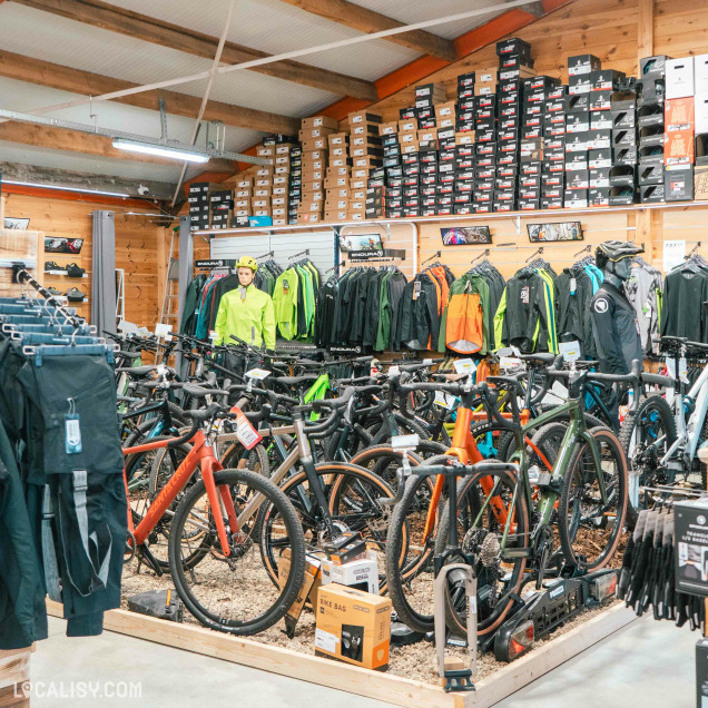 L'intérieur du magasin de vélos Ardennes Bike à Spa, avec une grande variété de vélos exposés au centre, entourés de vêtements de cyclisme accrochés aux murs et de nombreux cartons de chaussures empilés sur des étagères en hauteur. Les vêtements incluent des vestes et des pantalons de différentes couleurs, principalement des teintes vives comme le vert et l'orange. Il y a également des accessoires de vélo comme des casques et des sacs.