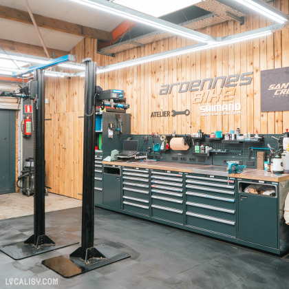 L'intérieur de l'atelier du magasin de vélos Ardennes Bike à Spa, avec un espace de travail bien organisé avec des outils et des équipements de réparation de vélos. Il y a des établis avec de nombreux tiroirs de rangement, des outils accrochés au mur, et des équipements spécialisés comme un support de réparation de vélos. Le mur en bois ajoute une touche chaleureuse à l'atelier.