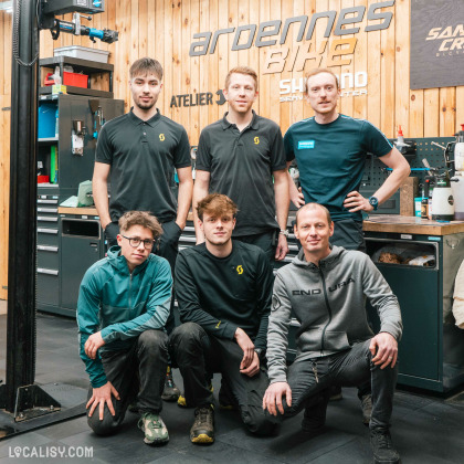 Un groupe de six personnes posant dans le magasin de vélos Ardennes Bike à Spa. L'atelier est bien équipé avec des outils et des équipements de réparation visibles en arrière-plan. Les personnes portent des vêtements de travail, certains avec des logos de marques de vélo. Le cadre en bois et les panneaux muraux affichent des marques comme Shimano et Santa Cruz Bicycles, ce qui indique que l'atelier est spécialisé dans ces marques.