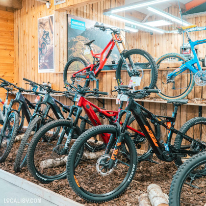 Plusieurs vélos de montagne sont exposés dans le magasin de vélos Ardennes Bike à Spa, certains accrochés au mur et d'autres posés sur le sol recouvert de copeaux de bois. Les vélos sont de différentes couleurs, notamment rouge, bleu et noir. Des affiches promotionnelles et des panneaux de marques comme Shimano sont visibles sur les murs en bois. L'éclairage est fourni par des lampes suspendues au plafond.