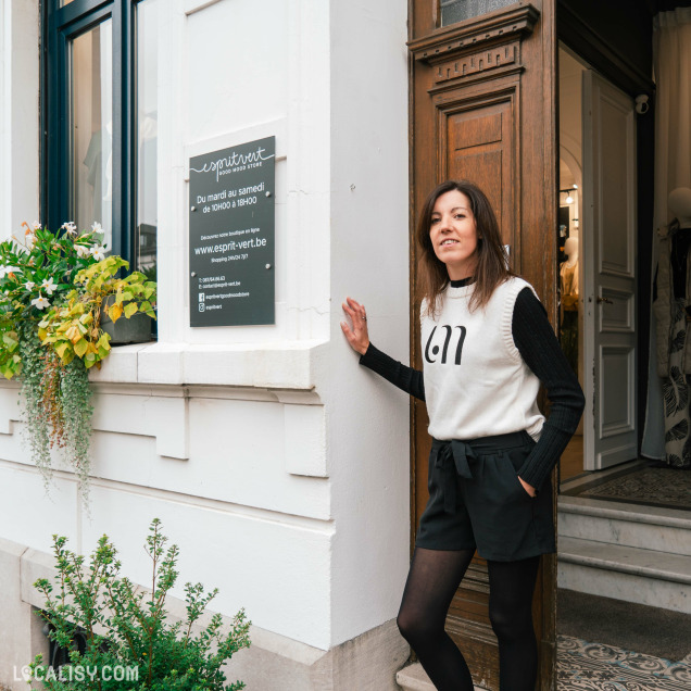 Devanture du magasin de vêtements pour femmes Esprit Vert à Heusy. Une personne debout à l'entrée du magasin Esprit Vert, la personne porte un pull blanc avec le numéro 611 et un short noir. À côté de la porte, il y a une enseigne noire avec des informations sur le magasin, y compris les heures d'ouverture (du mardi au samedi, de 10h00 à 18h00), le site web (www.esprit-vert.be), et les coordonnées de contact.