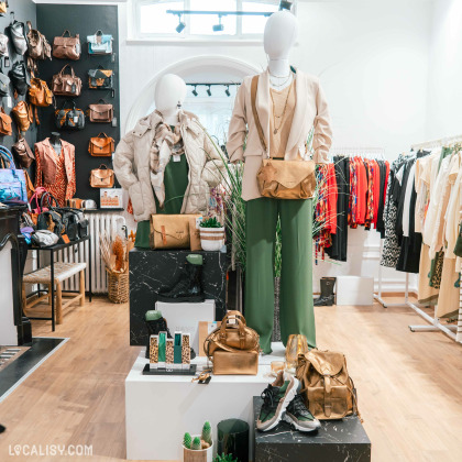 Deux mannequins habillés de vêtements élégants, l'un portant un pantalon vert et une veste beige avec un sac à bandoulière doré, et l'autre portant une veste matelassée beige avec une écharpe dans le magasin de vêtements pour femmes Esprit Vert à Heusy. Autour des mannequins, il y a divers sacs à main, sacs à dos, chaussures et autres accessoires exposés sur des présentoirs. À l'arrière-plan, des étagères et des portants sont remplis de vêtements colorés et de sacs à main.