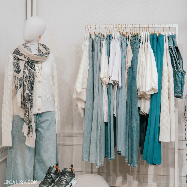 Un présentoir de vêtements dans le magasin de vêtements pour femmes Esprit Vert à Heusy. À gauche, un mannequin porte un cardigan blanc avec des motifs en relief, un jean large et une écharpe à motifs noirs et blancs. Le mannequin est également accessoirisé avec des colliers dorés. À droite, un portant de vêtements contient divers articles, principalement des jeans et des hauts dans des tons de bleu et de blanc. Au premier plan, une paire de baskets grises est posée sur un tabouret.