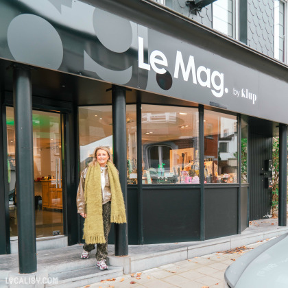 Devanture du magasin Le Mag by KLUP à Heusy – Façade moderne avec une enseigne noire et blanche élégante. Une personne souriante se tient devant l'entrée vitrée, laissant entrevoir un intérieur chaleureux et tendance.