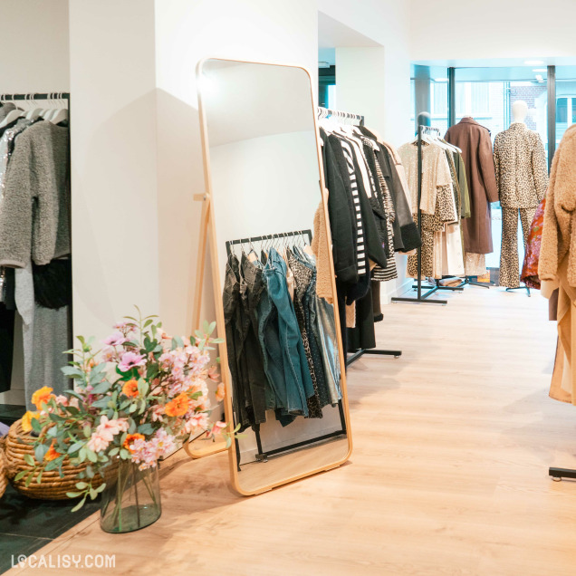 Rayons de vêtements et miroir décoratif – Espace lumineux avec des portants de vêtements élégants et un grand miroir en bois. Un vase rempli de fleurs colorées ajoute une touche de fraîcheur à l’ambiance.