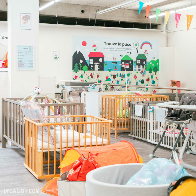 Espace mobilier et accessoires pour bébé – Zone colorée avec des lits pour bébé, des jouets et des équipements pour enfants. Une ambiance ludique avec un mur décoré d’illustrations et de drapeaux colorés.