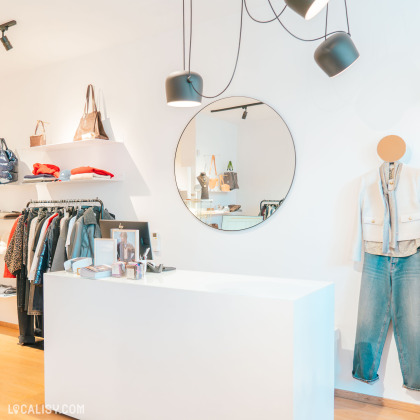 Espace d'accueil du magasin Syssi à Battice – Comptoir blanc épuré avec un miroir rond moderne. Des vêtements et accessoires élégants sont exposés sur des étagères et des portants.