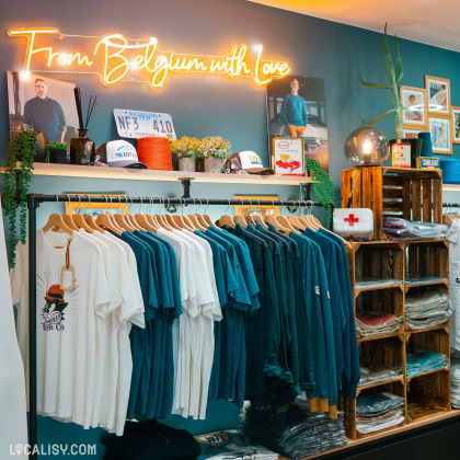 Intérieur de la boutique avec un mur bleu décoré d'un néon lumineux affichant "From Belgium with Love", des vêtements pliés et suspendus, et une ambiance chaleureuse.