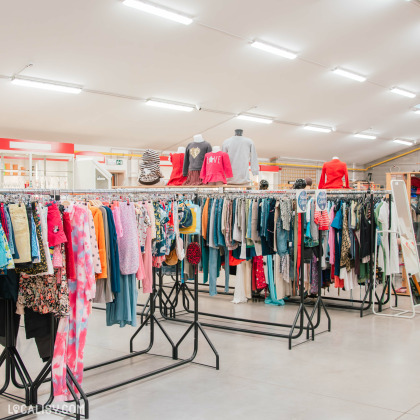 Intérieur du magasin de seconde main 3R second hand à Lontzen, avec des vêtements variés exposés sur des portants.