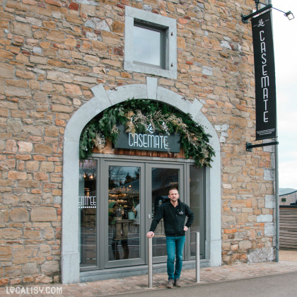 Façade en pierre du magasin d'alimentation La Casemate à Theux avec son propriétaire devant l'entrée.