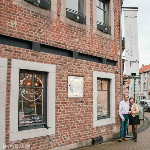 Façade en briques rouges de la boutique "Mam's Elles Foulards" avec de grandes fenêtres affichant le logo du magasin. Deux personnes se tiennent devant l'entrée, souriantes. Une bannière verticale et une enseigne dorée complètent l'esthétique élégante du magasin.