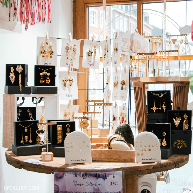 Présentation d'un stand de bijoux dans une boutique, avec des boucles d'oreilles en acier inoxydable et en plaqué or exposées sur des supports en bois et des cartons suspendus. Un panneau indique une réduction de 30 %. L'ambiance est lumineuse et chaleureuse.