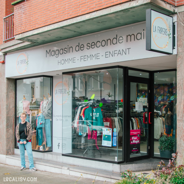 Façade du magasin de seconde main "La Friperie", avec une grande vitrine exposant des vêtements pour hommes, femmes et enfants. Une personne se tient devant l’entrée, souriante. L’enseigne et la signalétique indiquent les horaires d’ouverture.