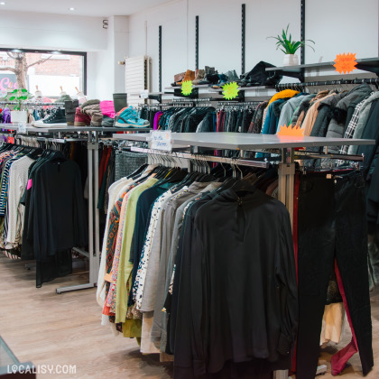 Intérieur de la boutique "La Friperie", avec une large sélection de vêtements suspendus sur des cintres. Des chaussures et accessoires sont exposés sur des étagères supérieures. Des étiquettes de prix et promotions sont visibles sur les articles.