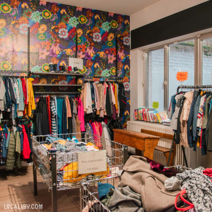 Coin dédié aux vêtements pour enfants dans la friperie, avec un mur coloré à motifs floraux. Des bacs remplis de vêtements pour bébés sont disposés au centre, avec une pancarte indiquant un prix à l’unité.