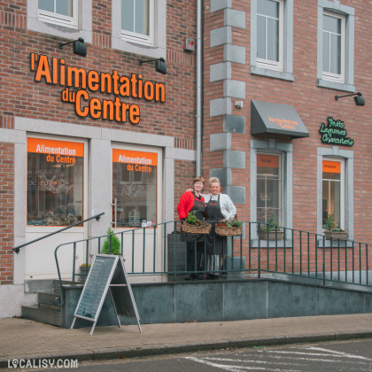 Façade du magasin "Alimentation du Centre", une épicerie locale avec une enseigne orange sur un bâtiment en briques. Deux commerçantes souriantes se tiennent devant l’entrée, portant des paniers de plantes. L'accès au magasin est facilité par une rampe, et une ardoise indique les produits disponibles.