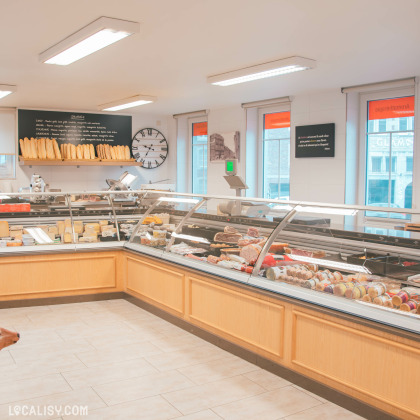 Intérieur de l'épicerie "Alimentation du Centre", présentant un vaste comptoir vitré rempli de fromages, charcuteries et produits frais. En arrière-plan, une étagère avec des baguettes bien alignées et une grande horloge murale décorent l’espace lumineux.