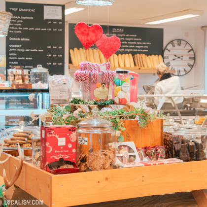 Présentation d’un étal festif dans l'épicerie "Alimentation du Centre", avec des produits gourmands comme des biscuits, chocolats et friandises emballées, accompagnés de décorations en forme de cœurs suspendus. Une ambiance chaleureuse et artisanale se dégage du présentoir en bois.