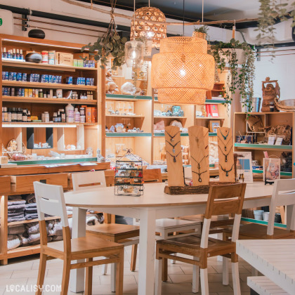 Espace boutique chaleureux avec une décoration en bois et un éclairage tamisé. Une table entourée de chaises blanches et en bois est installée au centre, tandis que des étagères exposent des bijoux, des cristaux et des produits de bien-être.