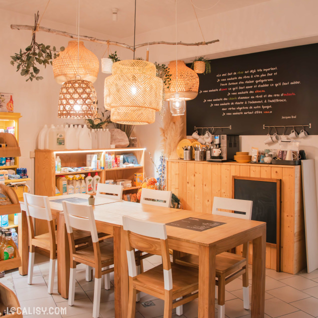 Coin détente dans une boutique bio avec une grande table en bois entourée de chaises. Des suspensions en rotin créent une atmosphère cosy, tandis qu’un mur noir affiche une citation inspirante. Des produits naturels et d’entretien sont soigneusement disposés sur des étagères éclairées.