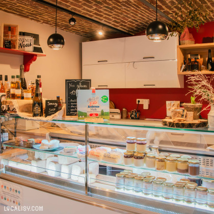 L’intérieur chaleureux du magasin La Cour Charlemagne, avec une vitrine remplie de fromages et de produits laitiers artisanaux. Derrière le comptoir, une décoration soignée avec des éléments en bois et des ardoises affichant les spécialités de la boutique.