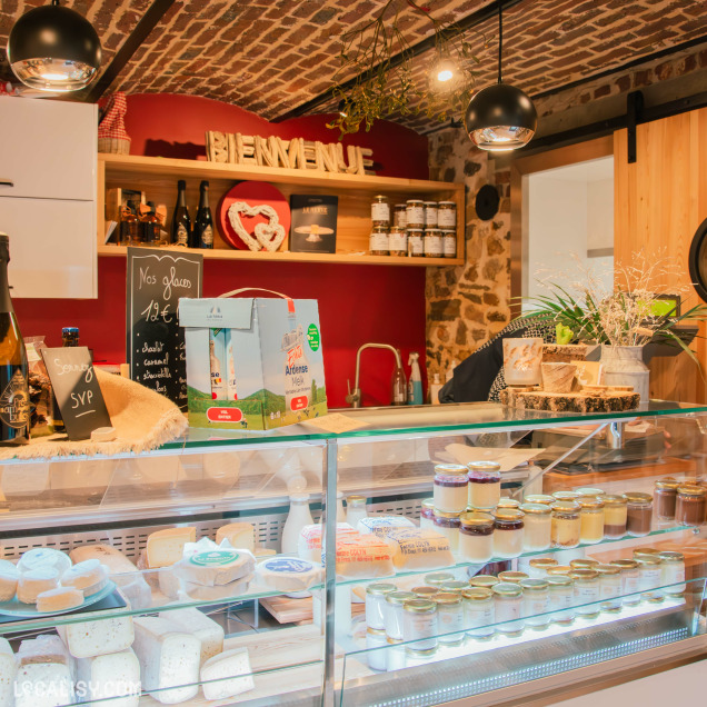 Une autre perspective du comptoir du magasin, mettant en valeur les fromages artisanaux et les produits laitiers sous une belle lumière. Des éléments décoratifs rustiques et un panneau "Bienvenue" ajoutent une touche chaleureuse à l’ambiance du commerce.