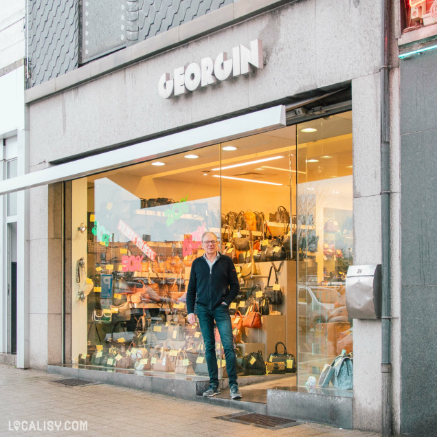 Façade du magasin "Georgin" avec un homme souriant debout devant l’entrée. La vitrine met en avant une grande sélection de sacs à main et de bagages.