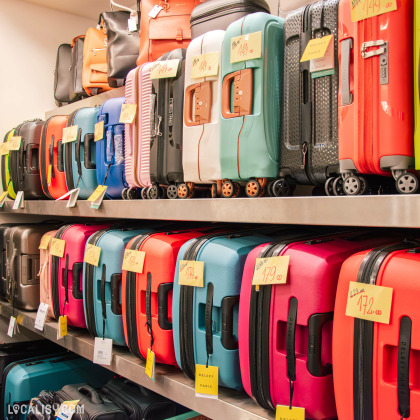 Rayons de valises colorées en exposition dans un magasin, avec des étiquettes de prix visibles. Les modèles varient en couleur et en taille, empilés sur des étagères.