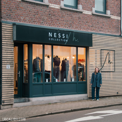 Façade de la boutique Nessi Collection à Battice, avec une devanture moderne et un logo en noir et blanc. Une femme souriante se tient devant l’entrée, et la vitrine met en avant des vêtements féminins tendance.